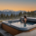 Spa extérieur haut de gamme avec une femme de dos savourant un verre face à un panorama de montagnes enneigées et de sapins au coucher du soleil. La couverture du jacuzzi est partiellement repliée sur le côté.